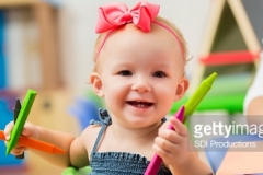 Cute little toddler girl sitting at a table and playing with crayons with her teacher in the background. She is holding the crayons and smiling at the camera.